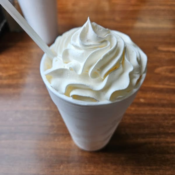 A white styrofoam cup of root beer float with whipped cream and a straw on a wooden table.