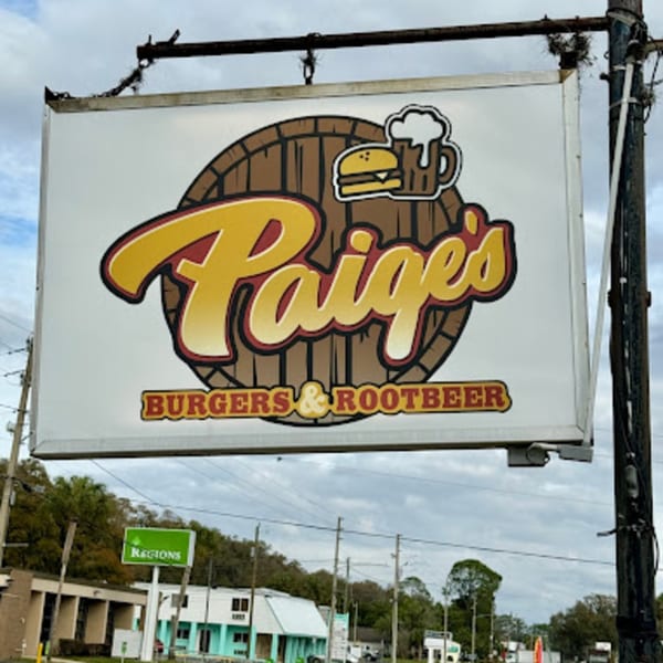 A white sign for Paige's Burgers & Rootbeer, featuring a burger, a root beer mug, and a barrel.