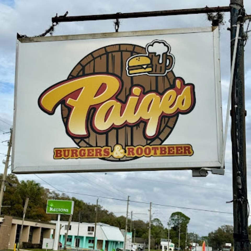 A white sign for Paige's Burgers & Rootbeer, featuring a burger, a root beer mug, and a barrel.