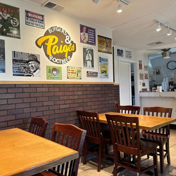 Interior of Paige's Root Beer restaurant with wooden tables, chairs, and vintage signs on brick and white walls.