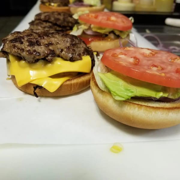 Several Paige's Root Beer cheeseburgers and hamburgers with fresh toppings on a white surface.