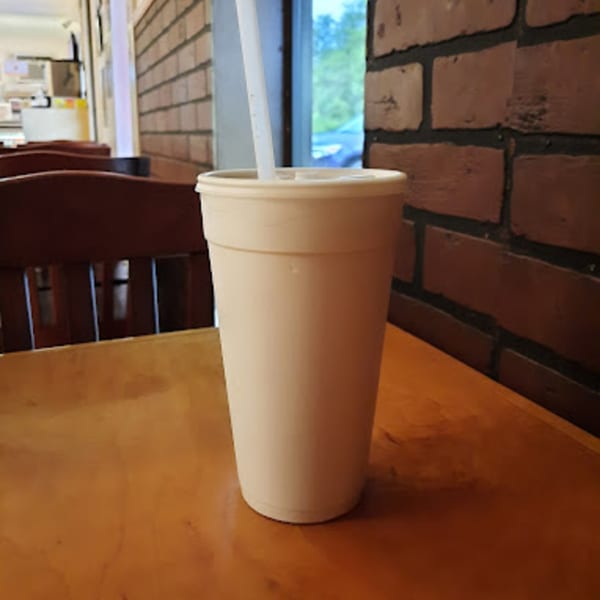 A Paige's Root Beer cup with a straw sits on a wooden table next to a brick wall.