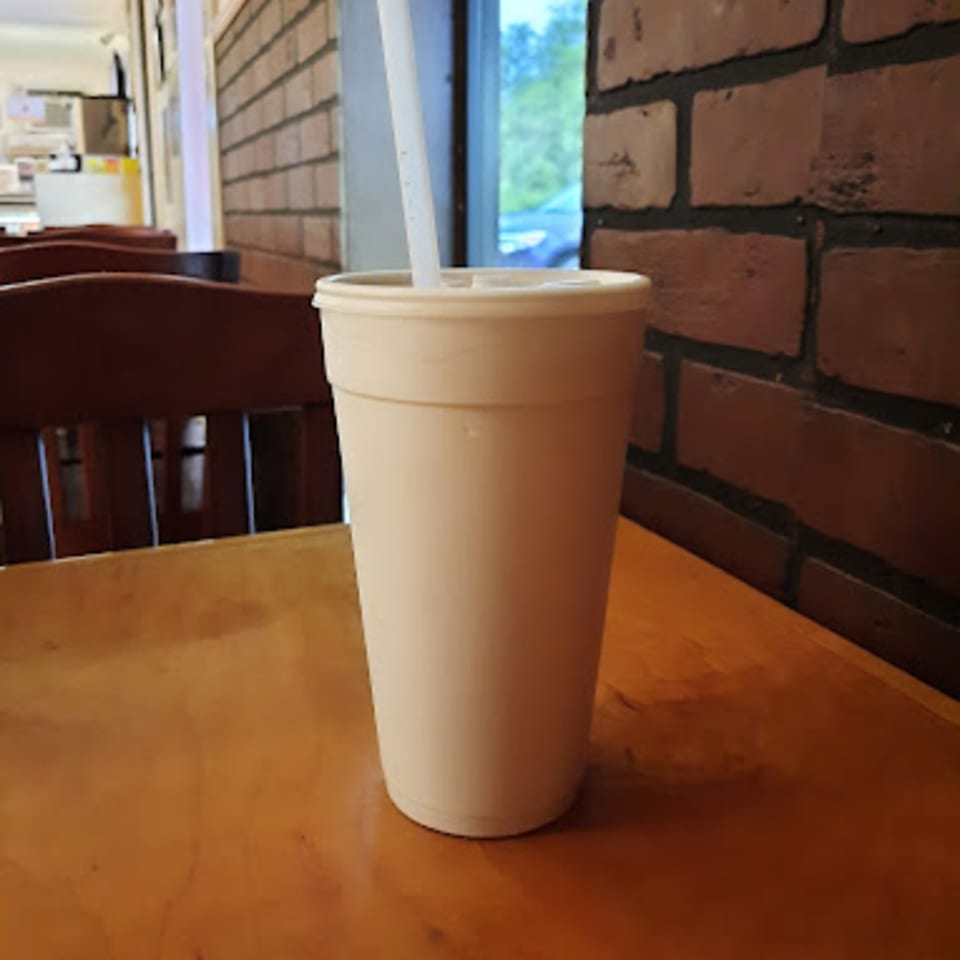 A Paige's Root Beer cup with a straw sits on a wooden table next to a brick wall.